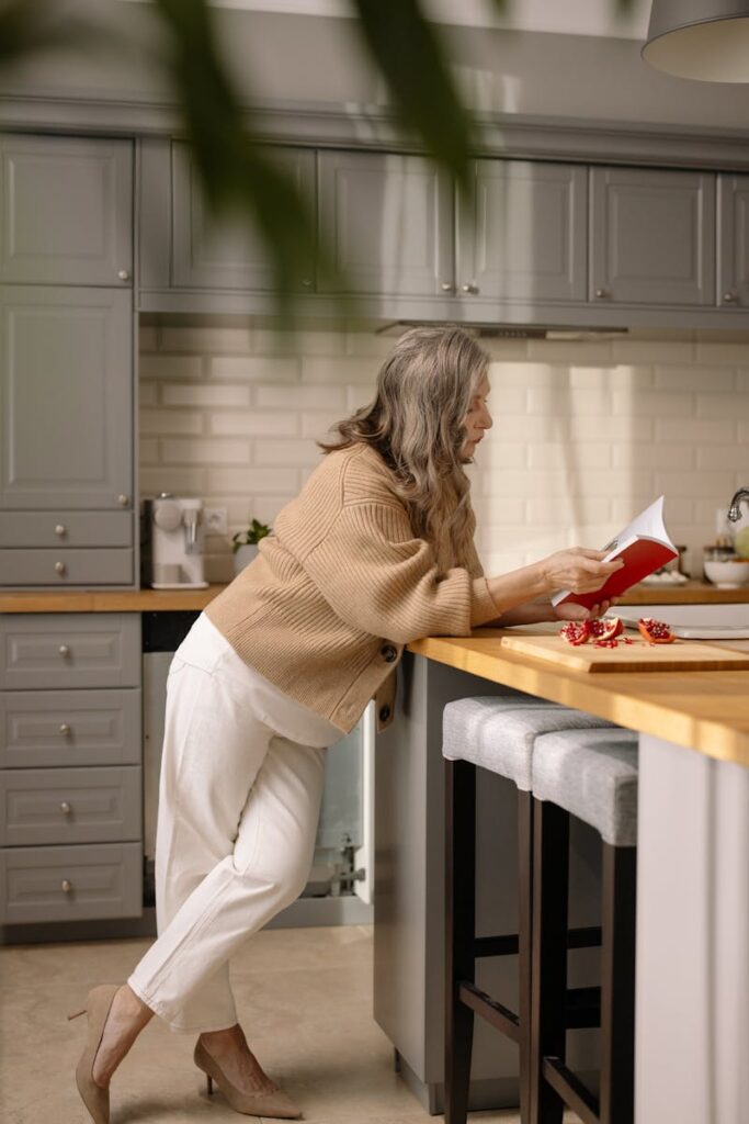 Elderly woman in kitchen reading a book, surrounded by a cozy atmosphere and fresh pomegranates.