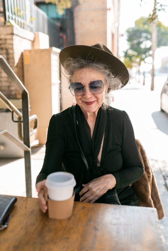 Senior woman in a hat and sunglasses enjoying coffee outside on a sunny day. Embracing life after 60 with style.