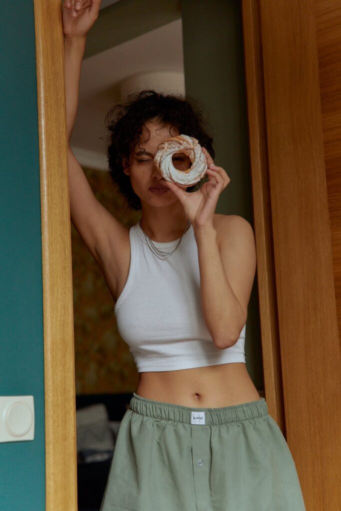 Fashionable woman standing in a doorway playfully holding a donut to her eye indoors.
