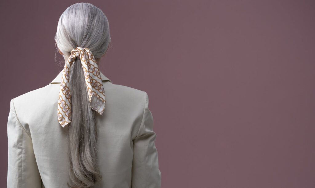 Back view of a stylish woman with a beige blazer, gray hair tied in a ponytail with a decorative scarf.