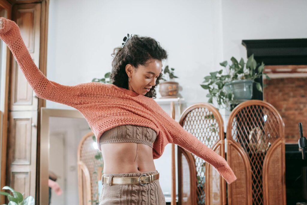 Positive African American female with dark curly hair wearing beige knitted top and leggings with golden belt putting on light orange sweater