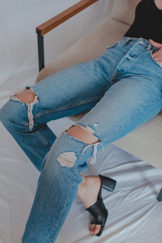 From above of anonymous woman in stylish ripped jeans and leather mule shoes sitting on chair placed on sheet