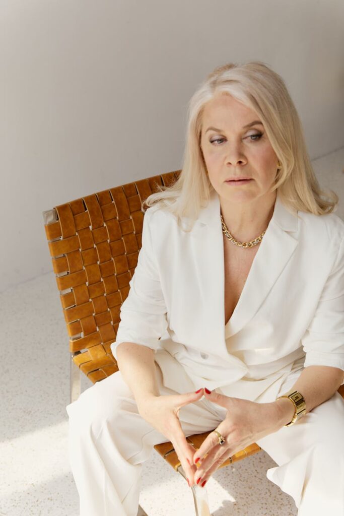 Sophisticated senior woman in a stylish white suit sitting on a modern chair indoors.