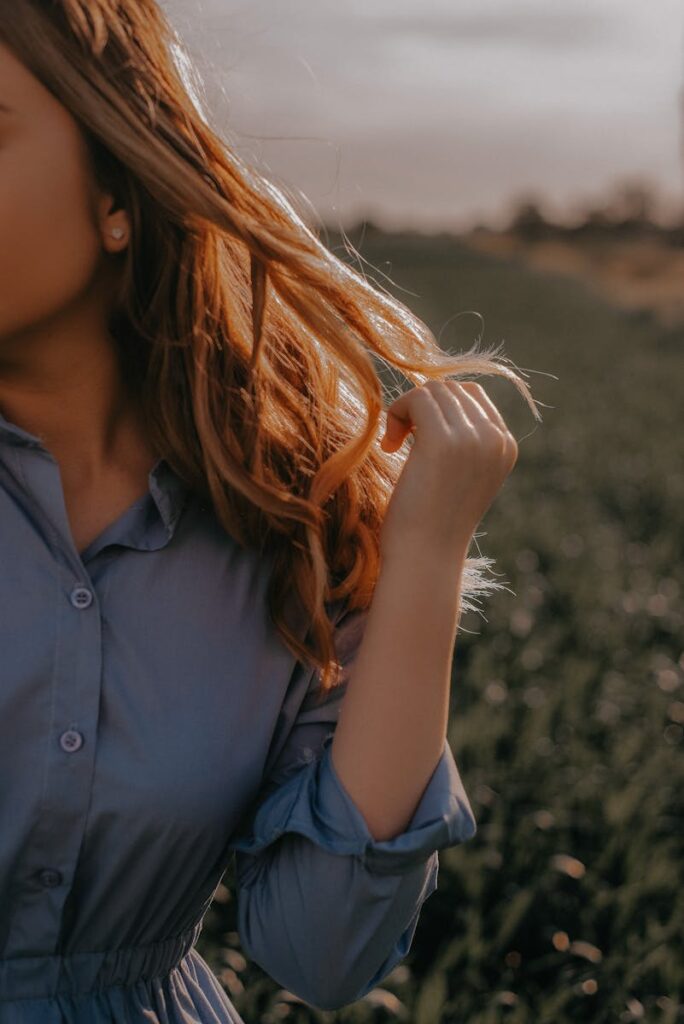 A woman gently holds her hair, wearing a blue dress in a sunlit outdoor setting.