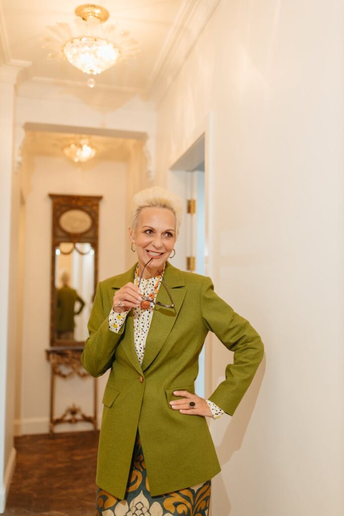 Portrait of a fashionable senior woman in a green blazer, standing confidently indoors with a drink.