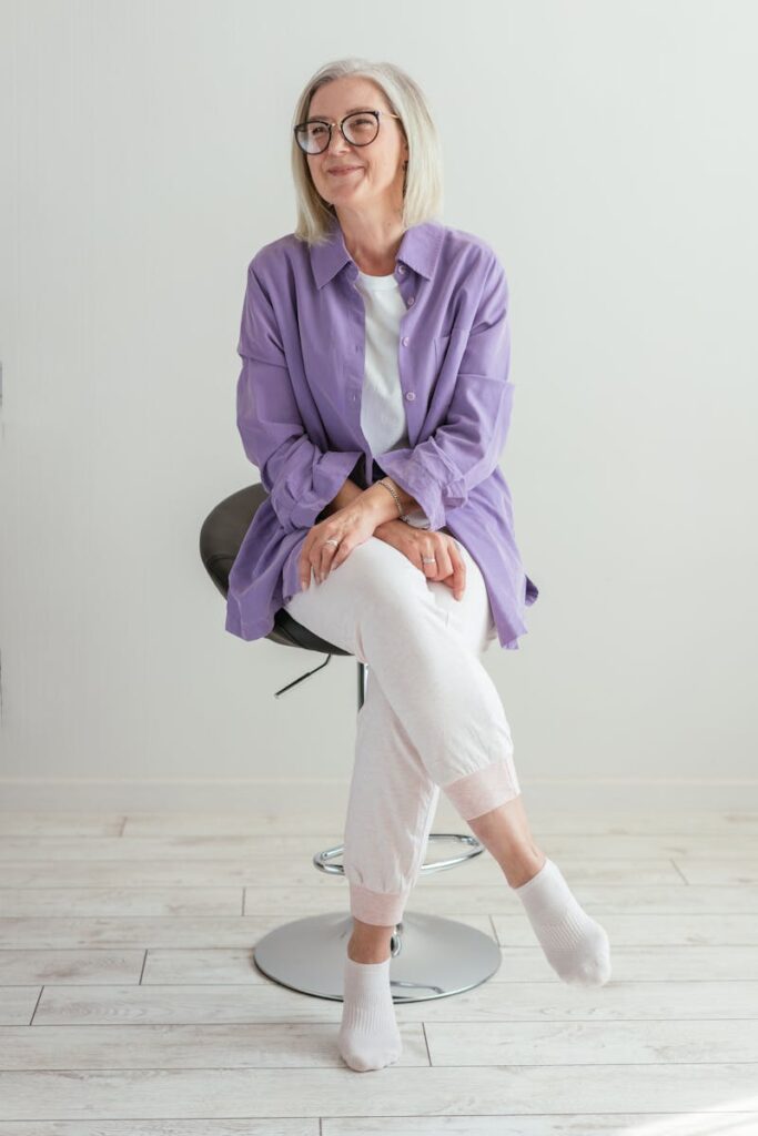 A senior woman in a purple shirt sitting on a stool and smiling in a bright studio.