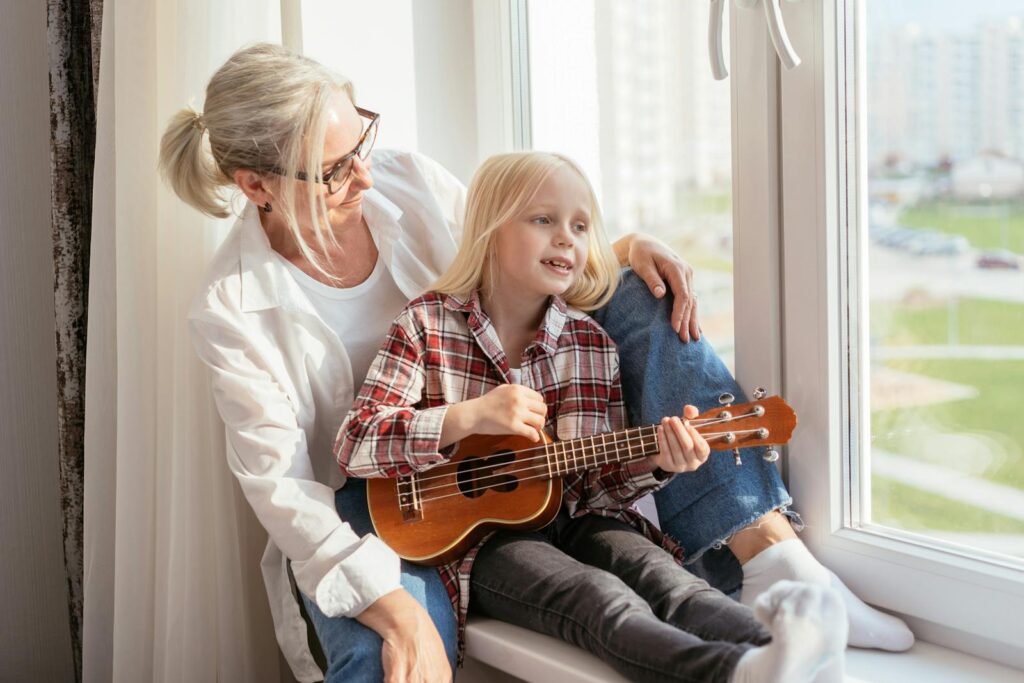 Grandmother and granddaughter sharing a joyful moment playing ukulele by the window.