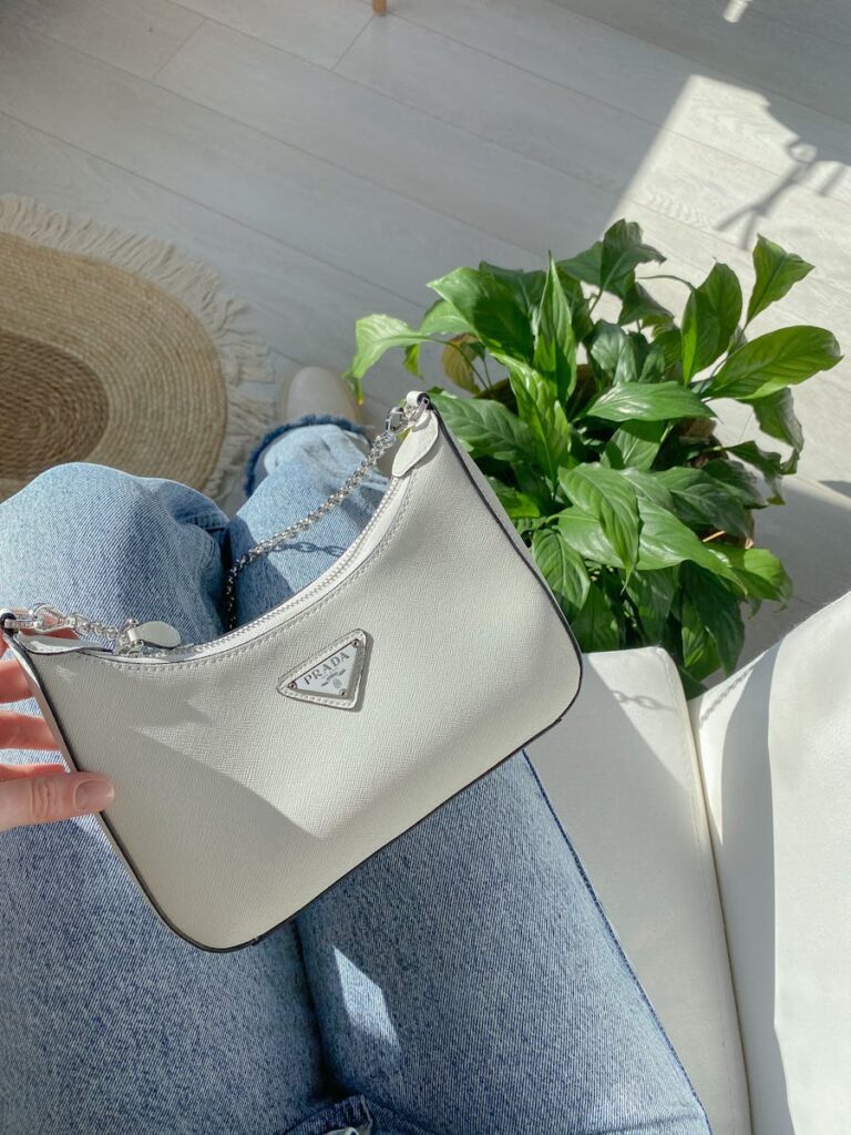 Stylish white designer handbag resting on denim jeans, surrounded by indoor sunlight and plants.