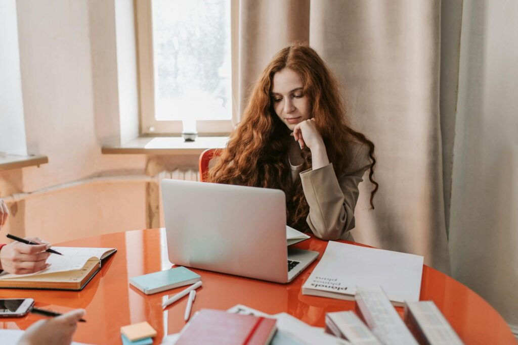 Redhead woman working on laptop in bright office setting with documents and notes on a red table.
