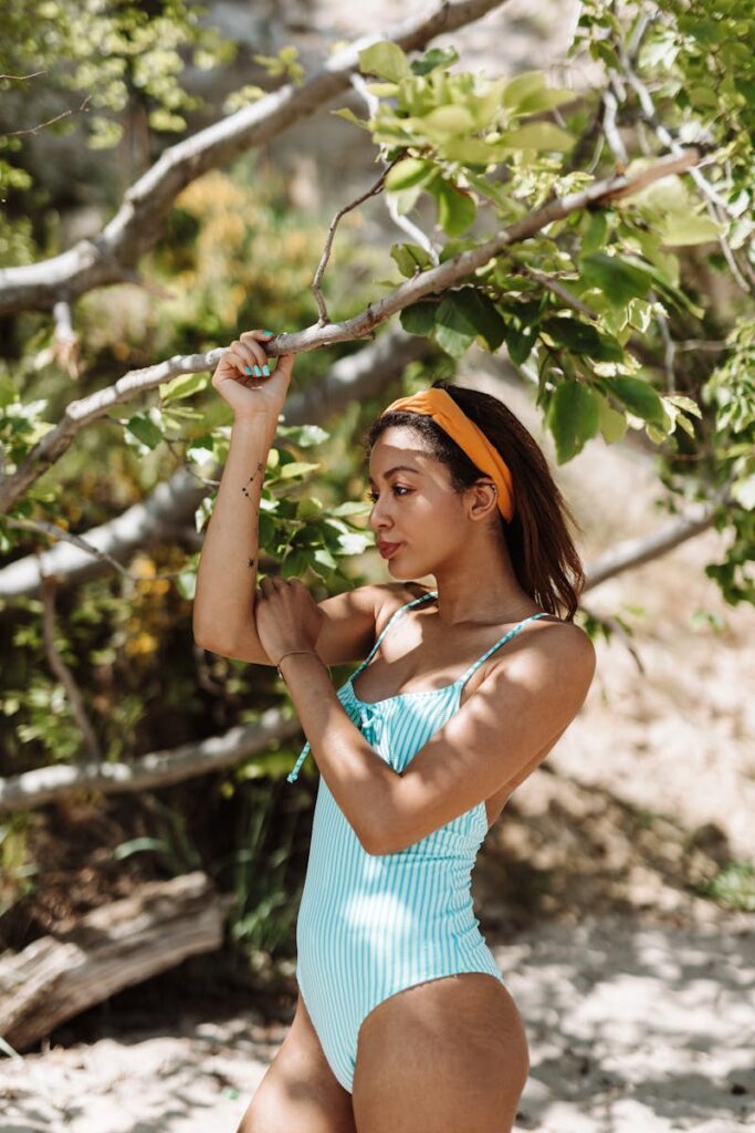 A young woman in a blue swimsuit poses under tree branches on a sunny day.
