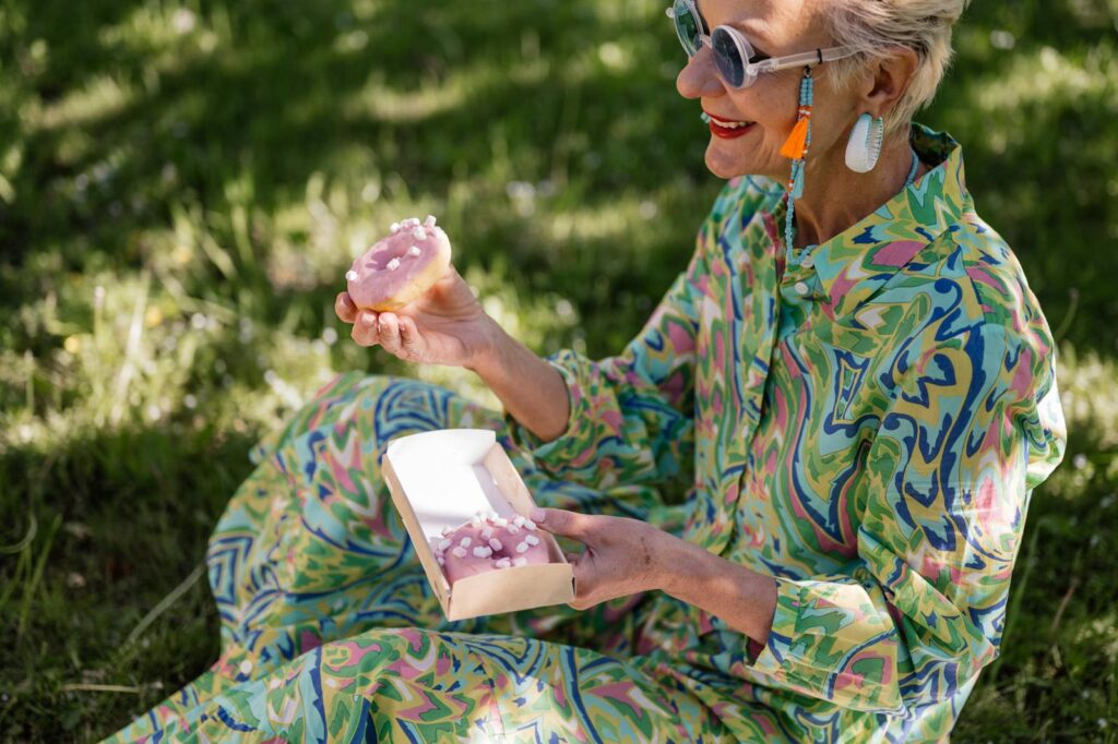 Senior woman in a colorful dress enjoying donuts outdoors during a sunny picnic.