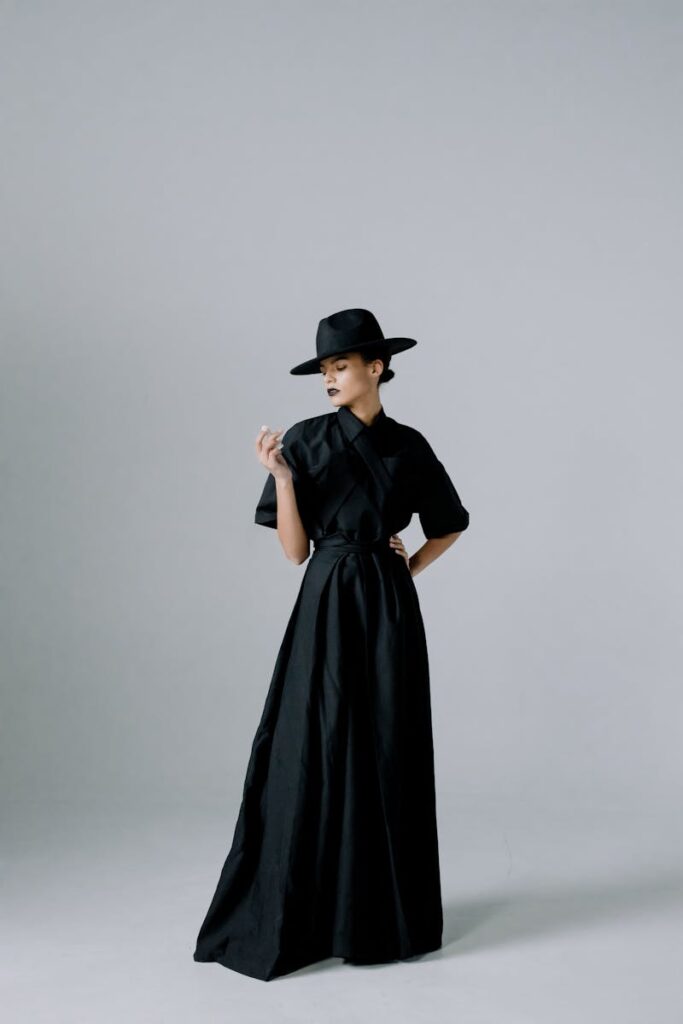 Stylish adult woman in a long black dress and hat posing confidently indoors.