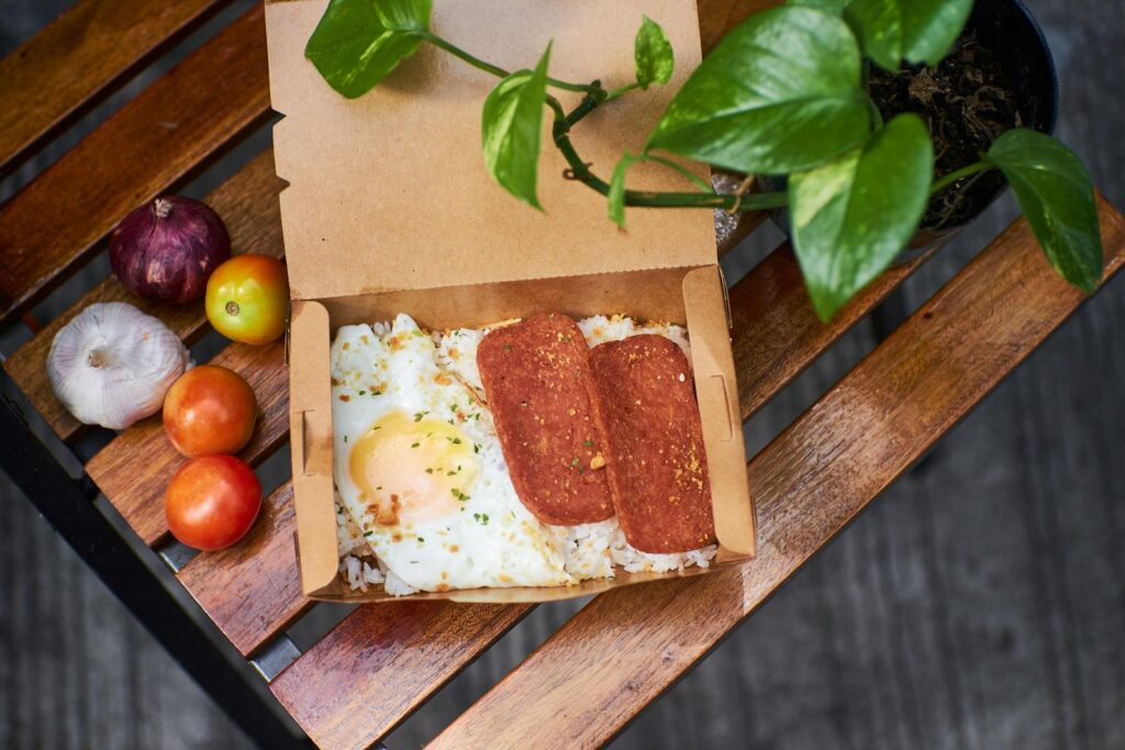 Delicious breakfast box featuring fried egg, sliced meat, and fresh vegetables on a rustic wooden table.
