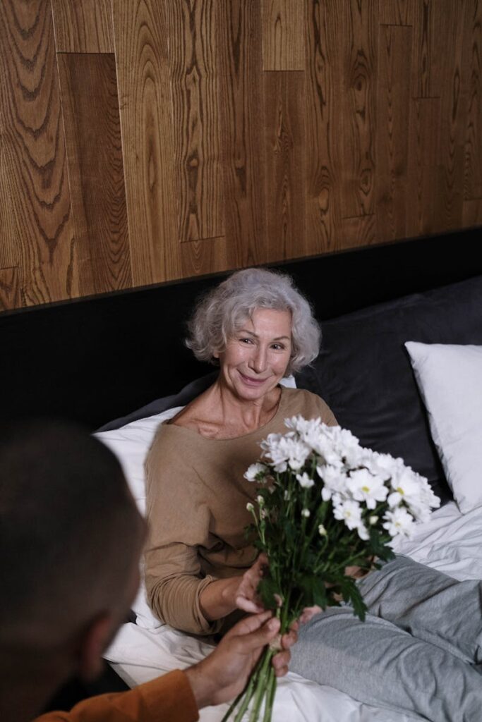 Senior woman smiling while receiving a bouquet of daisies in a cozy bedroom setting.