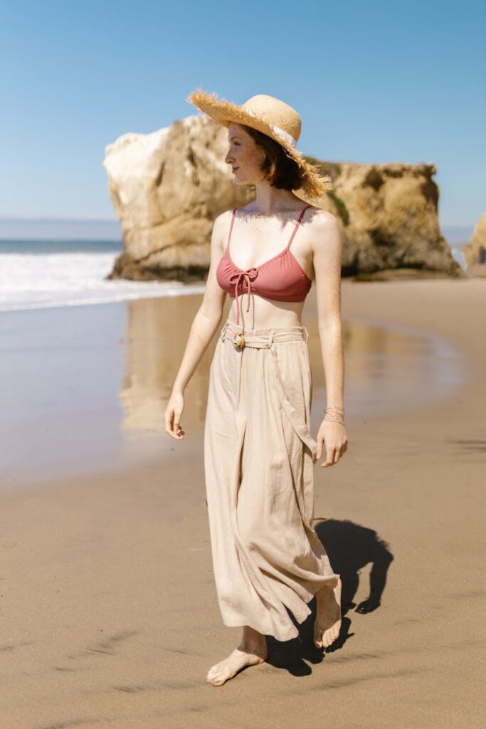 A woman in a straw hat and bikini top walks along a sunny beach shore.