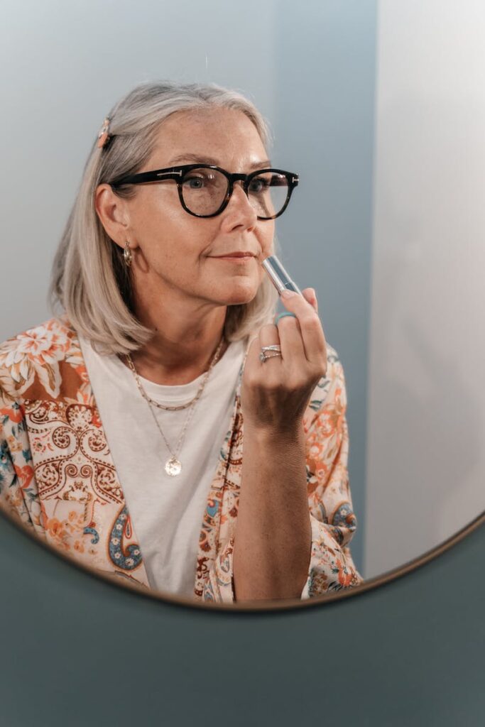 Senior woman in glasses applying lipstick, reflecting self-care and confidence.