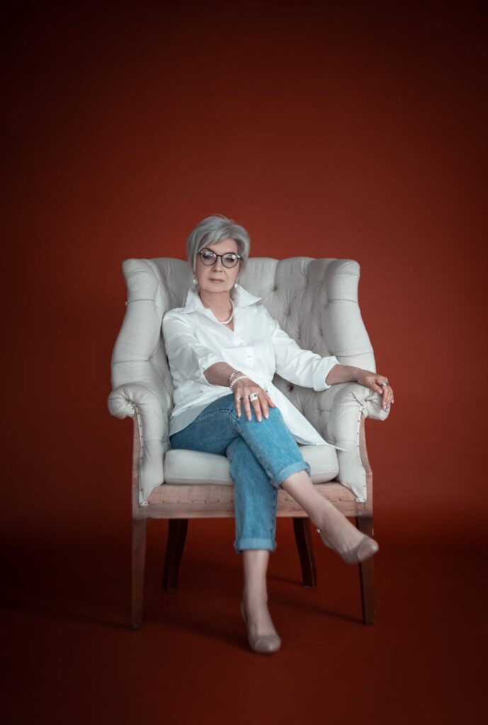 Stylish senior woman with grey hair and eyeglasses posing elegantly in an armchair indoors.