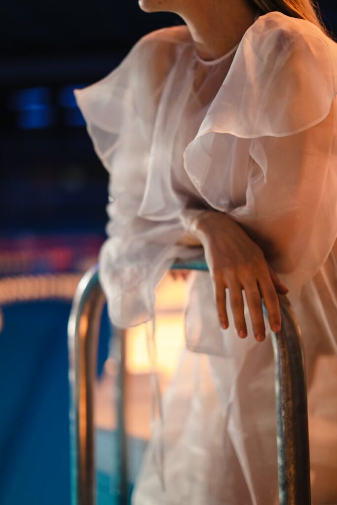 A woman in a delicate sheer dress leaning on a pool grab rail, creating an ethereal and moody atmosphere.