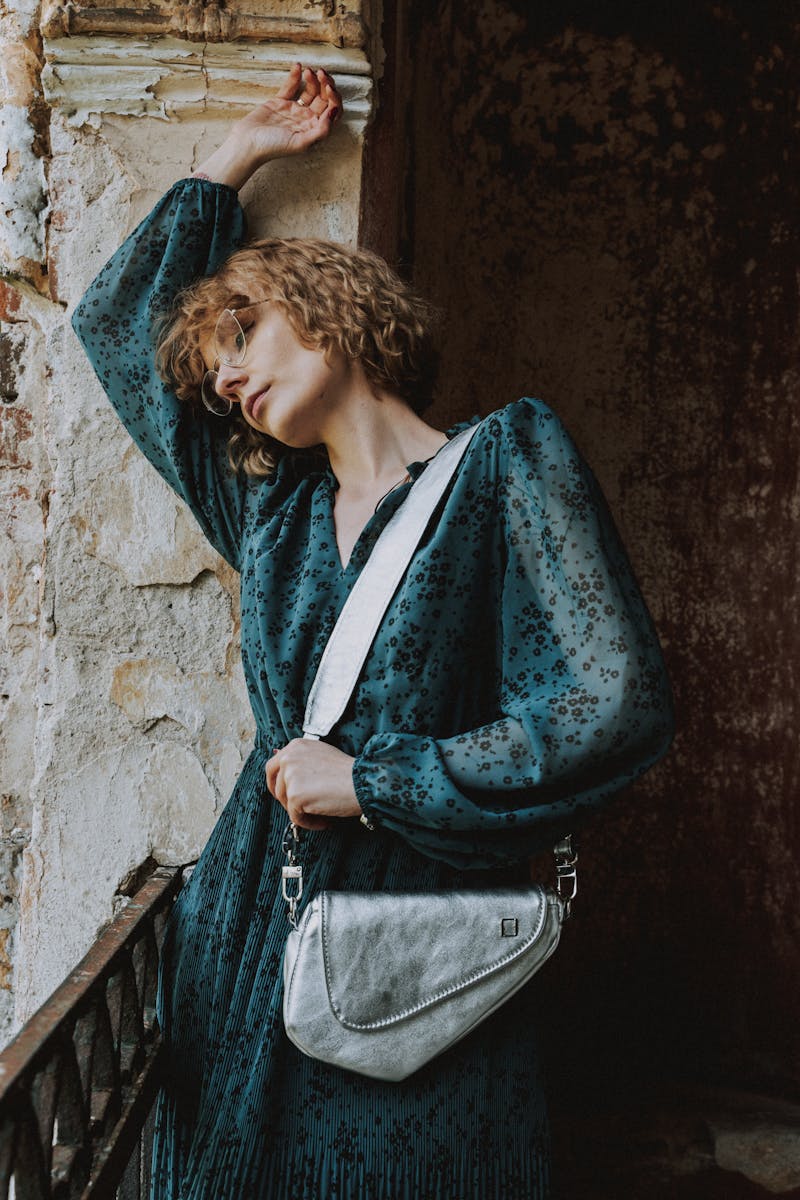 Elegant woman with short hair in a teal dress and eyeglasses, leaning thoughtfully against a rustic wall.