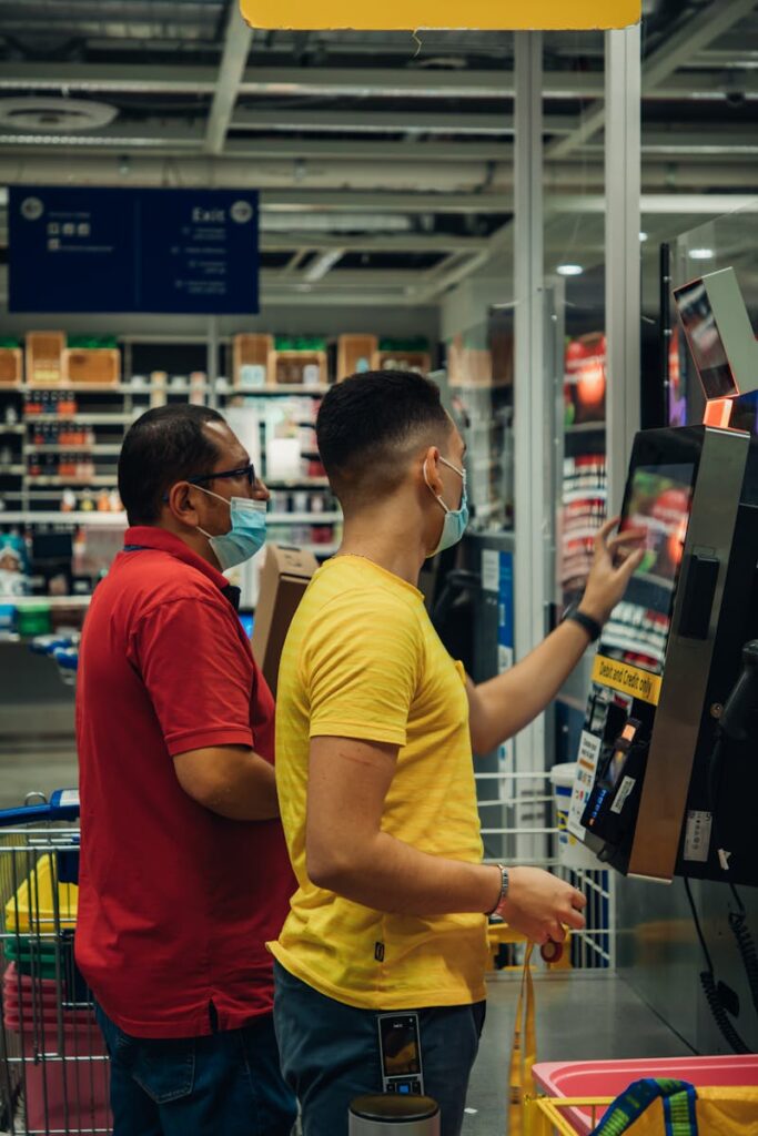 Two men wearing masks using a self-checkout machine in a grocery store.
