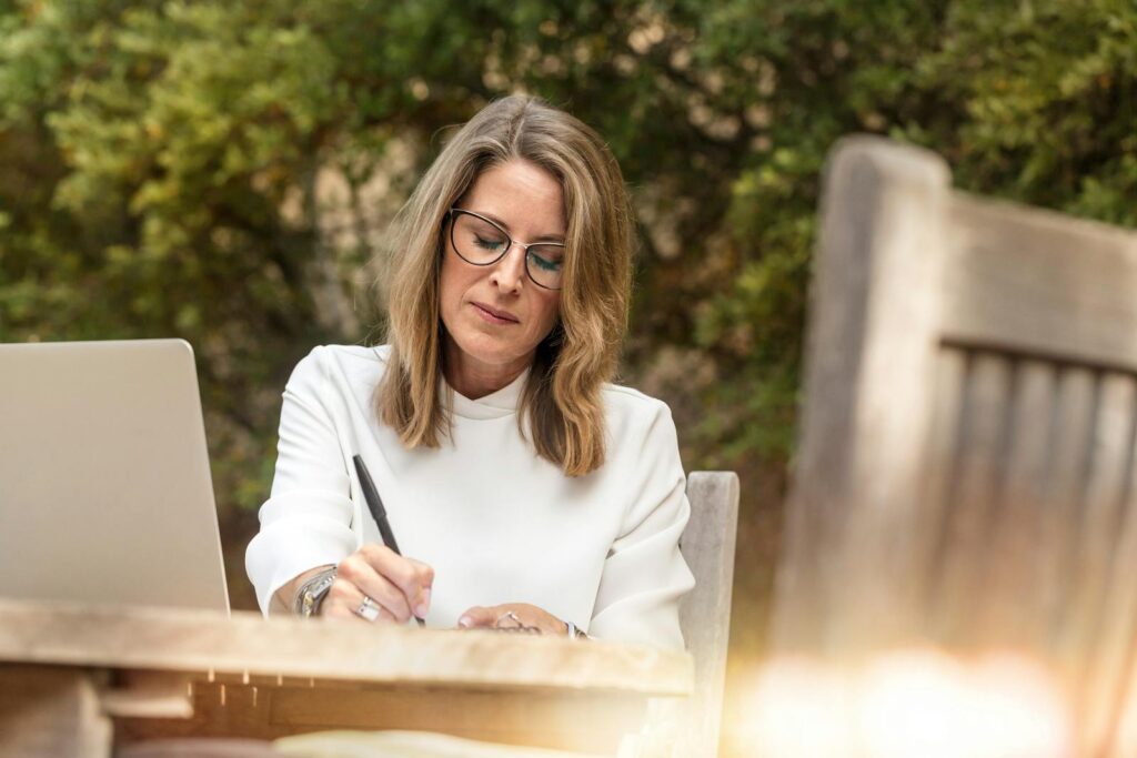 A woman in glasses writing and working on her laptop outdoors in natural daylight.