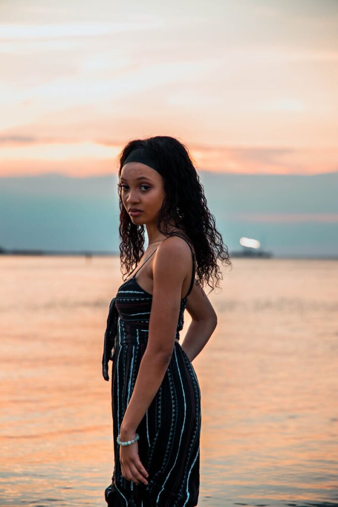 A woman stands by the sea during a tranquil summer sunset, exuding elegance and calm.