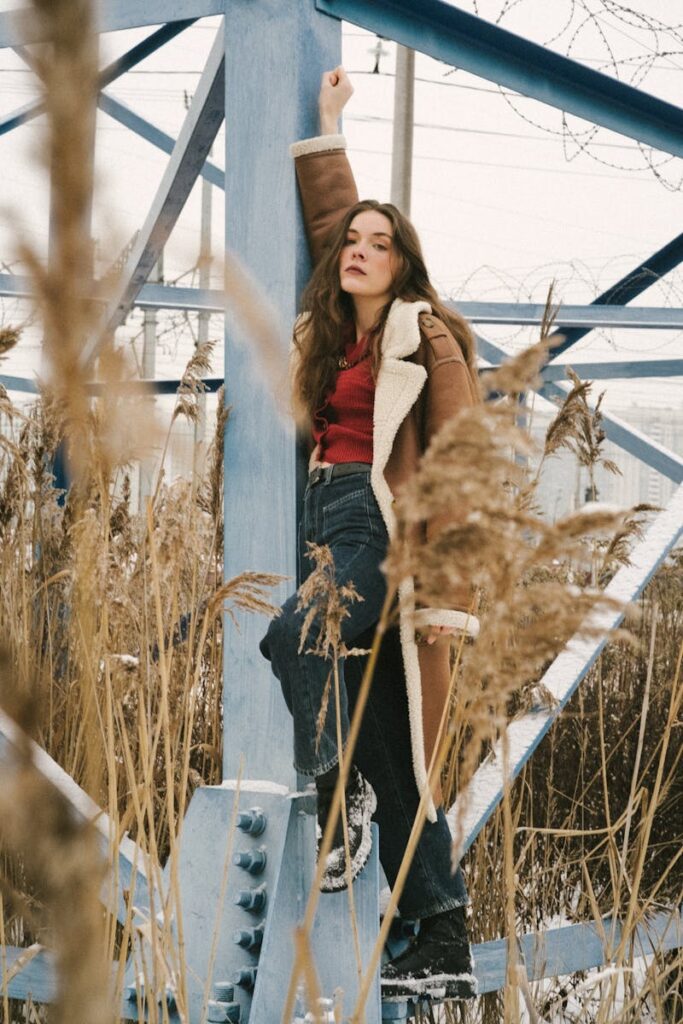 Woman posing in winter coat amidst industrial structures and dry grass.