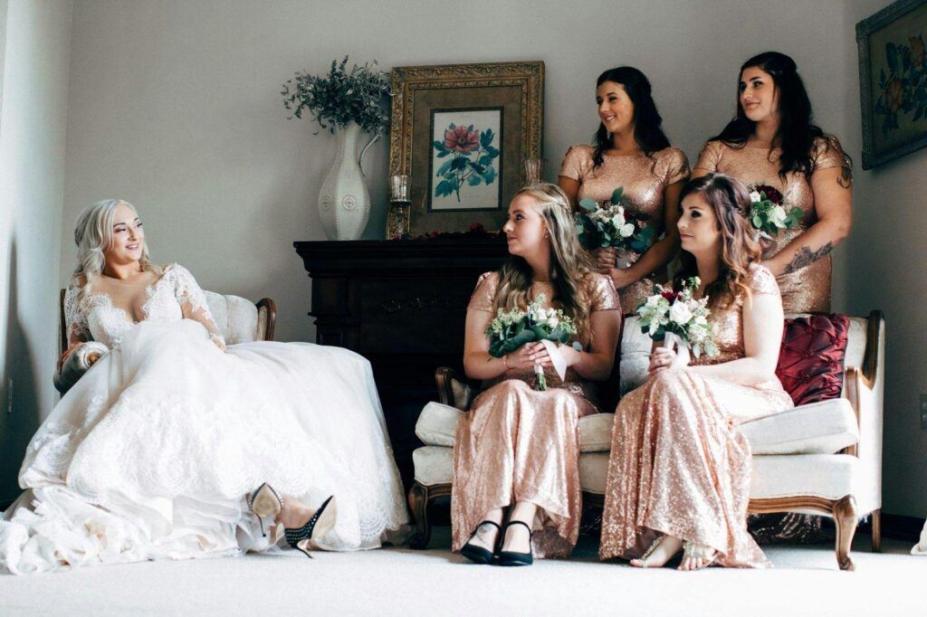 A bride in a lace dress and bridesmaids in sequin gowns share a candid moment indoors.