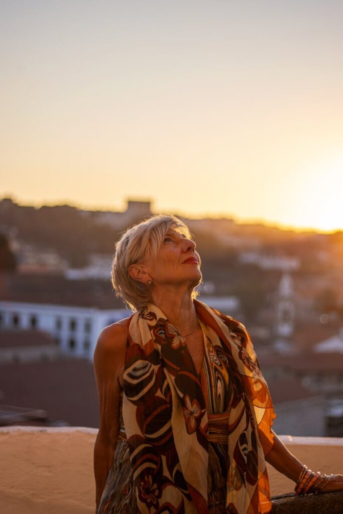 A senior woman in stylish attire looks up during a serene urban sunset, embracing tranquility.