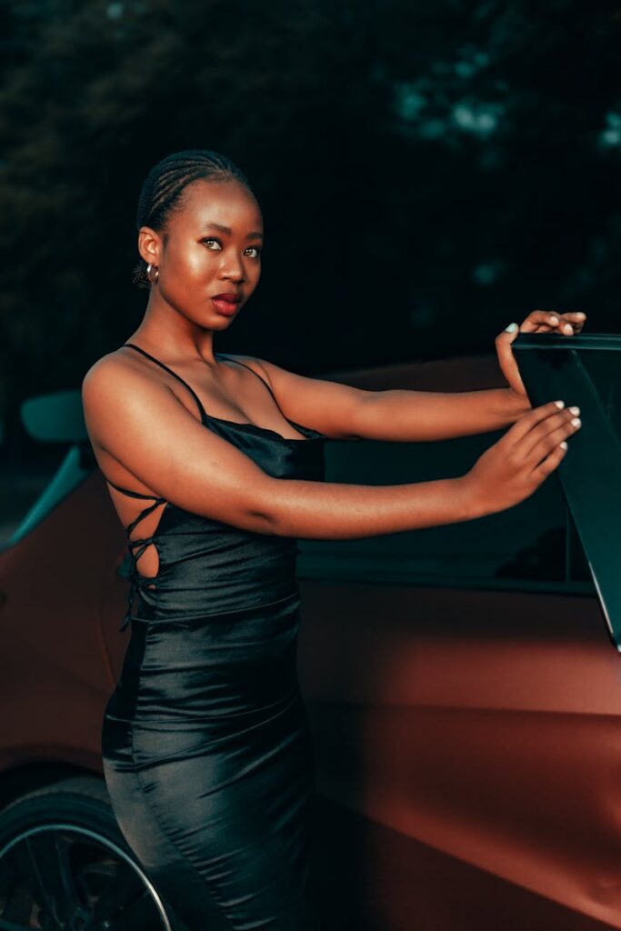 A fashionable woman in a black dress poses elegantly next to a car during twilight.