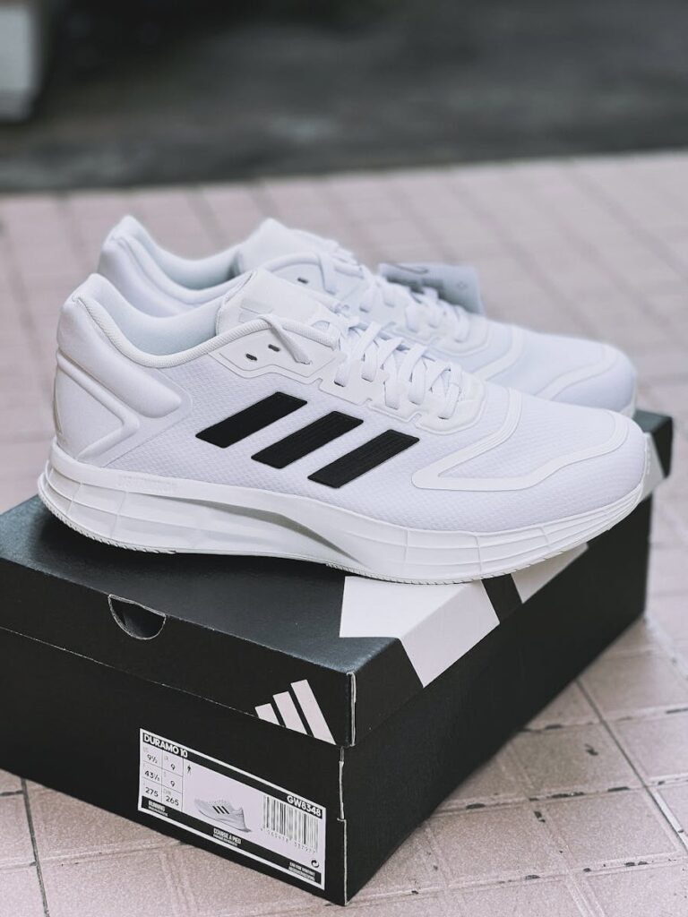 A pair of stylish white sneakers with prominent black stripes displayed on their box outdoors.