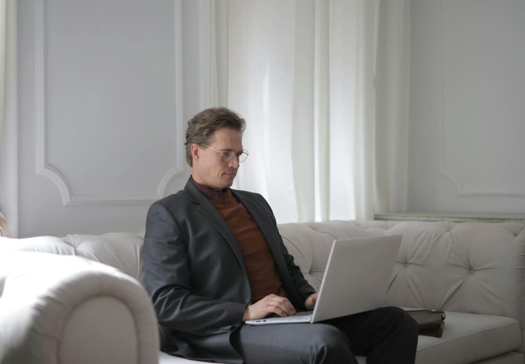 Professional man in glasses using a laptop while sitting on a white sofa at home office.