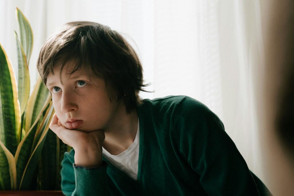 A child with a pensive expression sitting indoors next to a house plant.