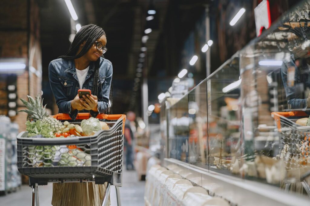 A young woman with afro braids grocery shopping in a supermarket, using a mobile phone.