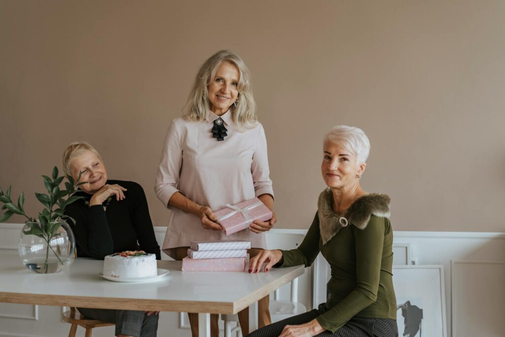 Three senior women enjoying a birthday party with gifts and cake indoors.