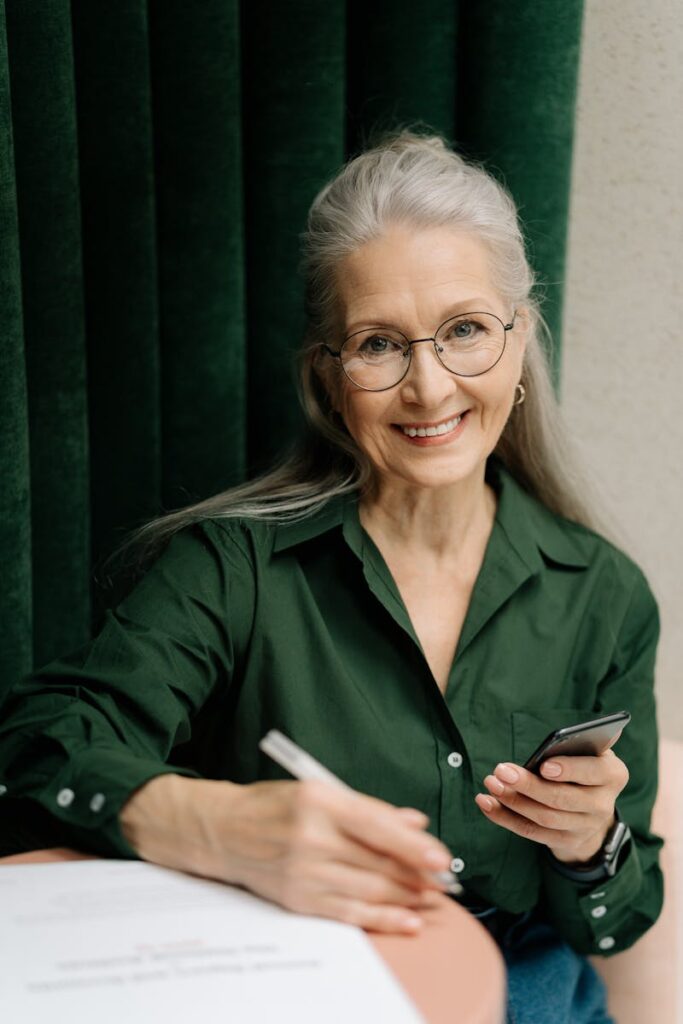 Smiling senior woman with gray hair and glasses holding a smartphone indoors.