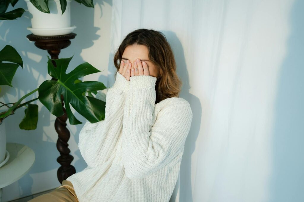 A woman in a white knitted sweater playfully covers her face with her hands indoors.
