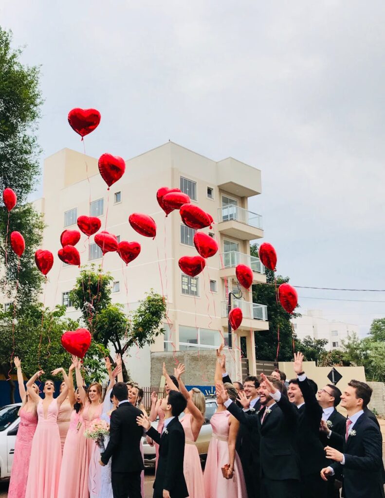A joyful wedding party celebrating outdoors with red heart balloons in the air against an urban backdrop.