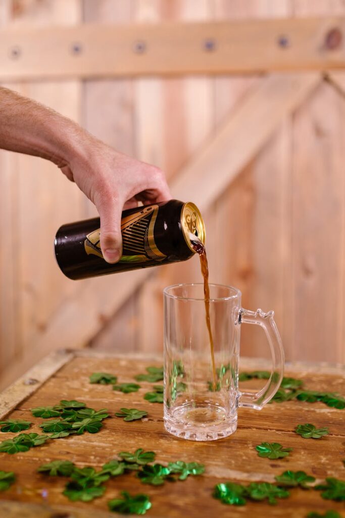 A hand pours dark beer into a glass on a wooden table decorated with green shamrocks.