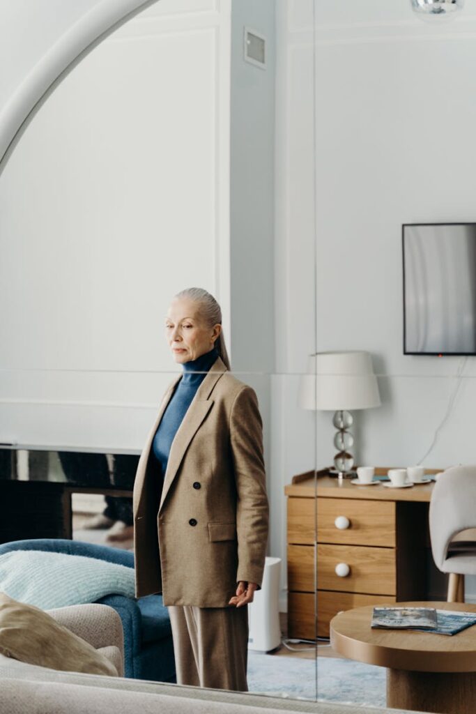 A stylish senior woman in a blazer stands indoors in a modern, elegant room with subtle decor.