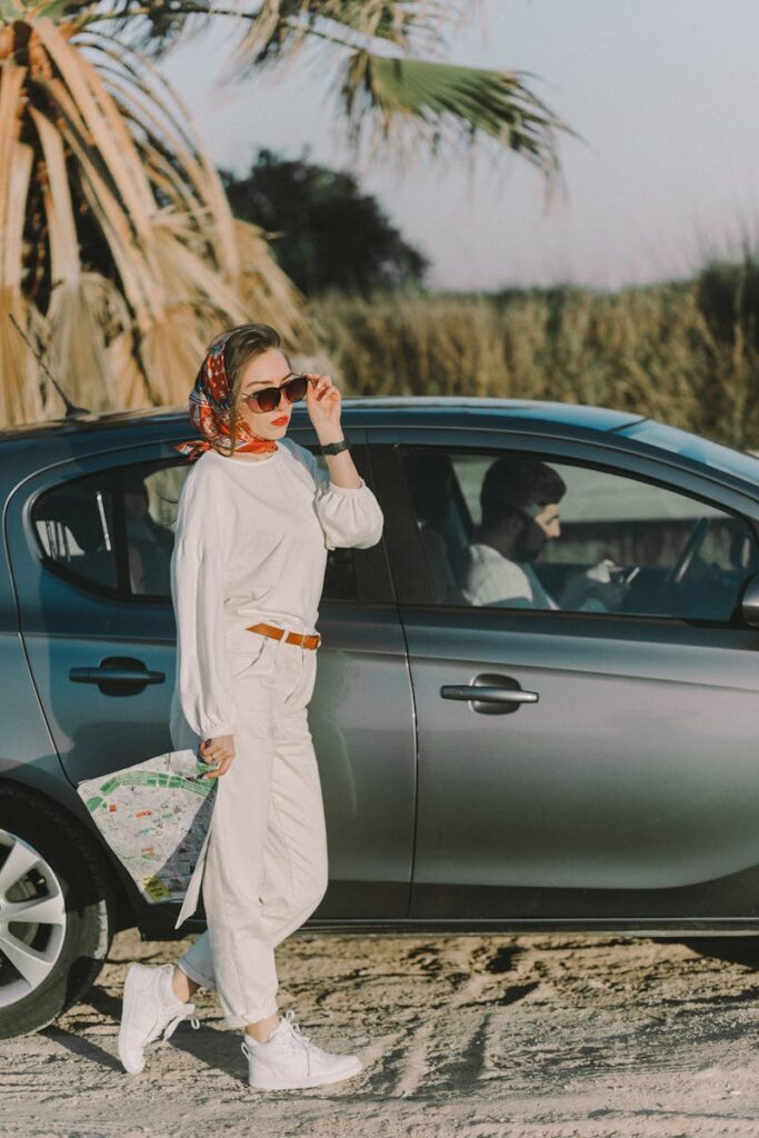 Stylish woman in sunglasses and scarf holding map beside parked car outdoors.
