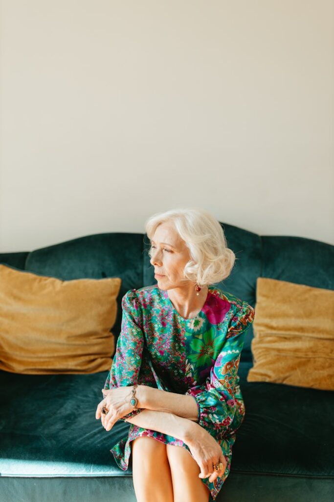 A stylish elderly woman in a colorful floral dress sits on a cozy sofa, embodying elegance and grace.