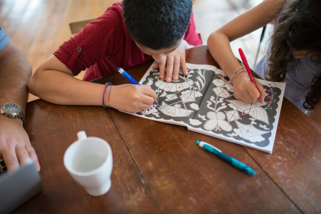 Two children coloring in an activity book with colorful pens on a wooden table.