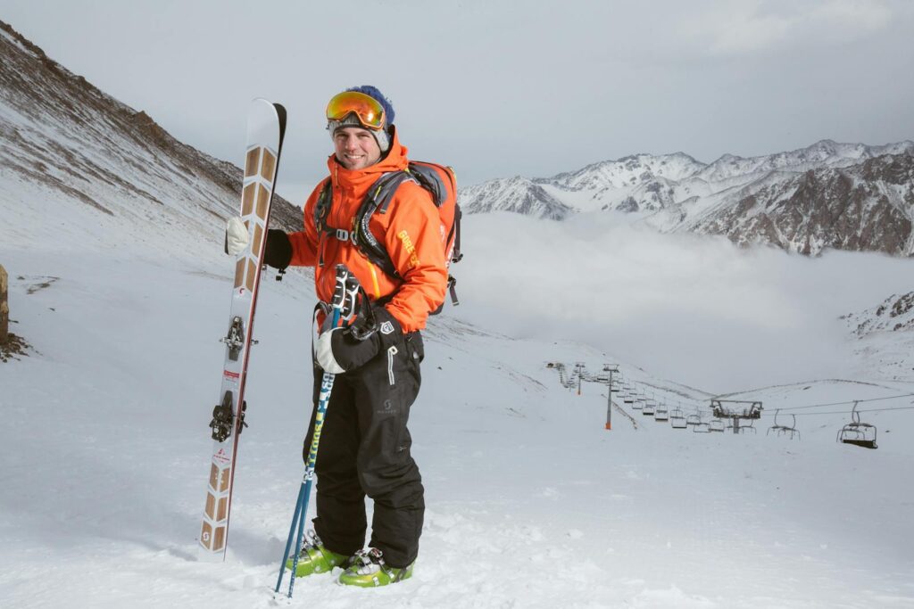 Smiling skier in orange jacket on snowy mountain with skis and poles ready for adventure.