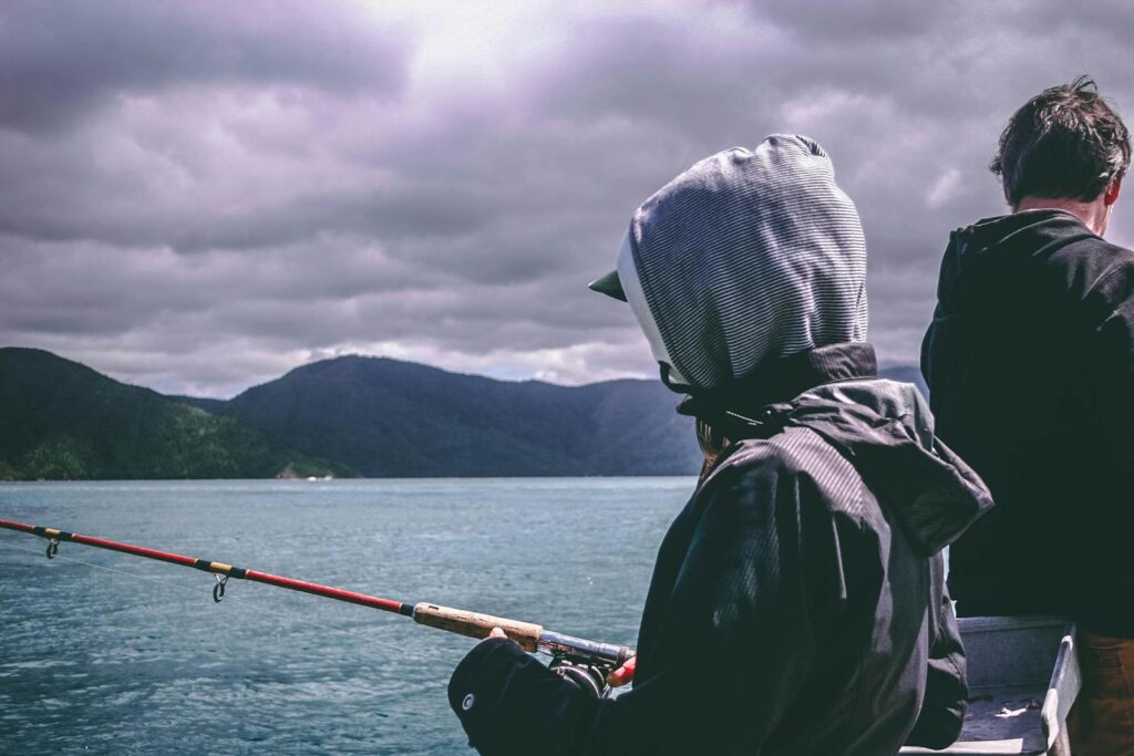 Two people fishing by the sea under a cloudy sky with mountain views.