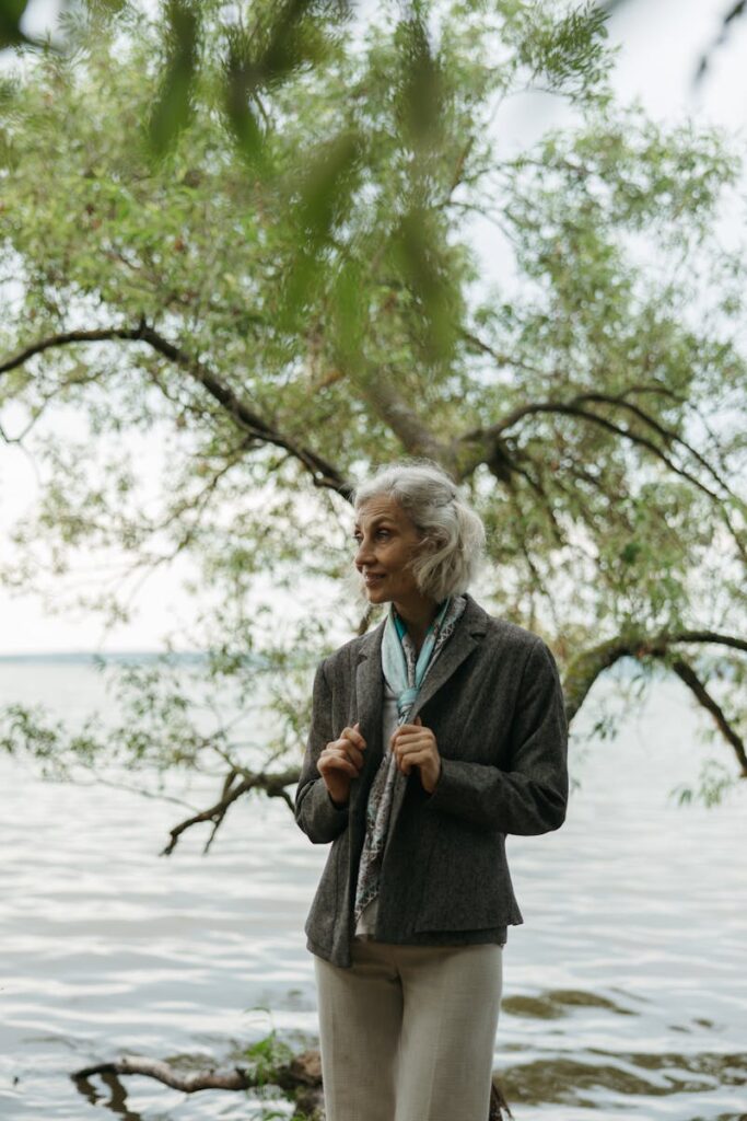 Senior woman standing by a lakeshore, surrounded by trees, enjoying the peaceful outdoor setting.