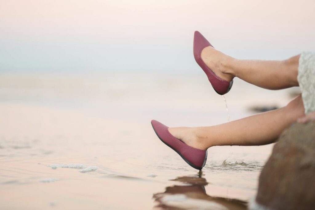 Women's legs wearing stylish maroon flats dipped in beach water, serene ambiance.