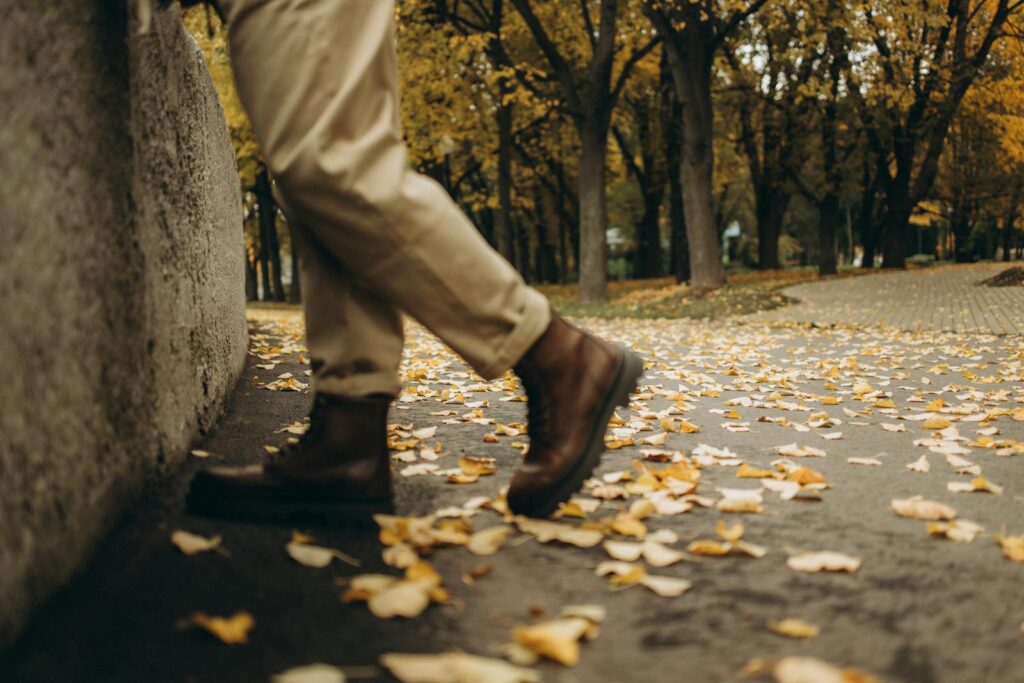 A person in boots walks through a park covered in autumn leaves.