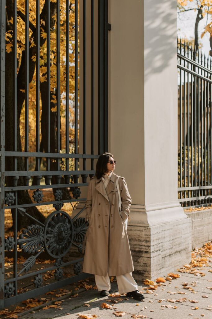 Woman in a beige trench coat leaning on ornate gate surrounded by autumn leaves.