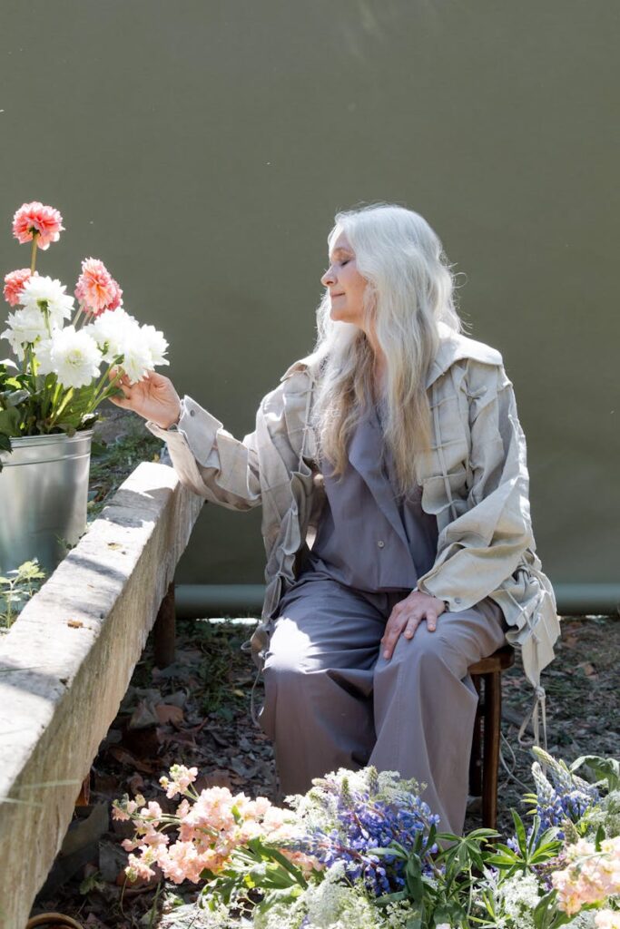 Elderly woman sitting peacefully surrounded by flowers in a greenhouse.